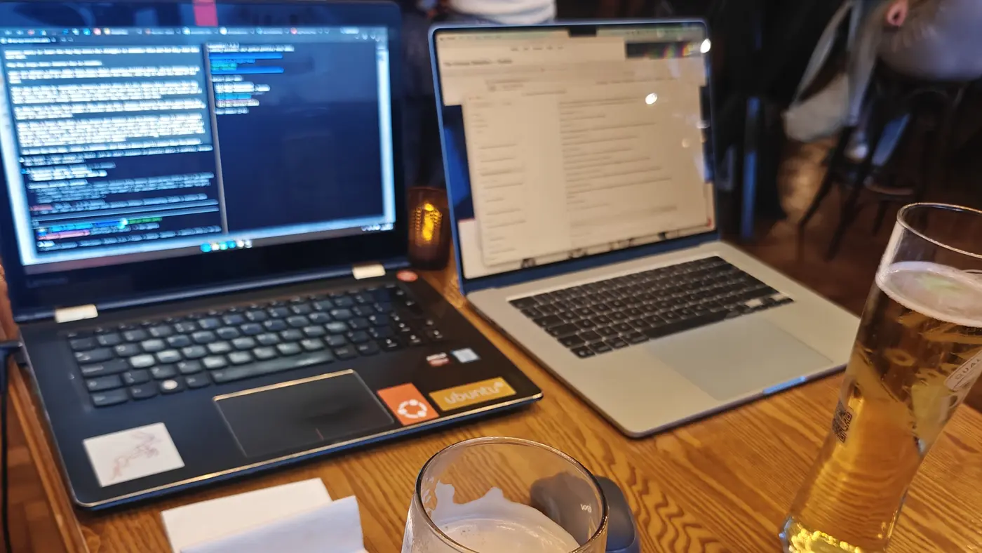 Two open laptops facing each other on a wooden pub table, one a Lenovo covered in Ubuntu stickers running a split-pane terminal session, the other a MacBook, with a half-drunk pint of lager and a stout glass between them and a busy pub behind.
