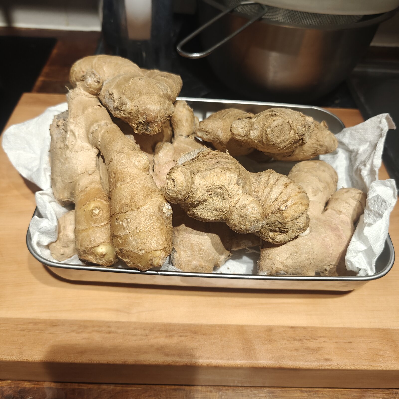 Tray of fresh ginger root knobs on kitchen paper, on a wooden chopping block, with a saucepan and strainer in the background. The 1kg I&rsquo;m still working through.